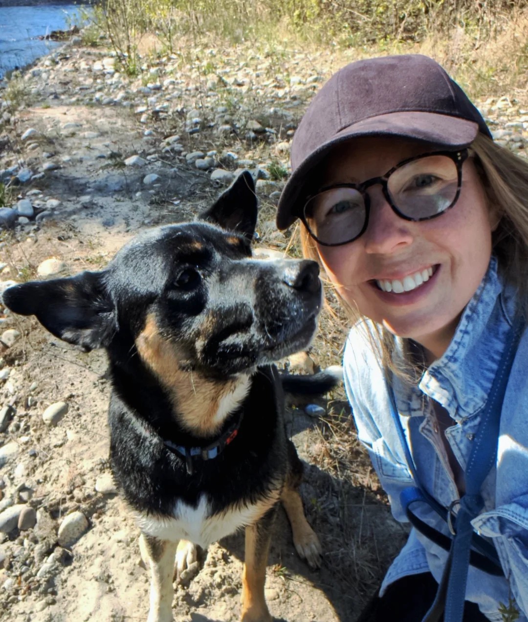 Breta Brassard smiling beside a black and tan cattle dog on a rocky riverbank